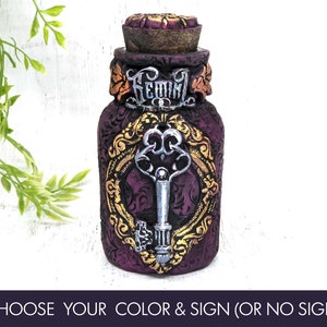 May include: A small purple glass bottle with a gold and silver key design. The bottle has a cork stopper and the word "Gemini" written on the front.