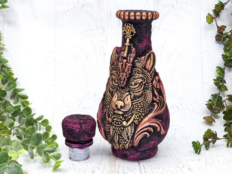 May include: A purple and gold ceramic bottle stopper with a wolf's head design. The stopper has a detailed design with intricate patterns and a gold finish.