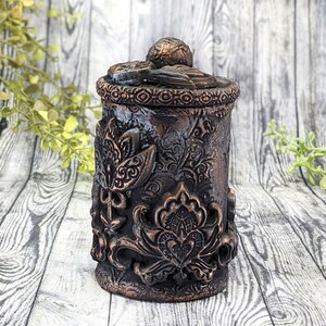 May include: A small, round, decorative container with a lid. The container is made of a dark brown material and has a textured, floral design. The lid is also decorated with a floral design.