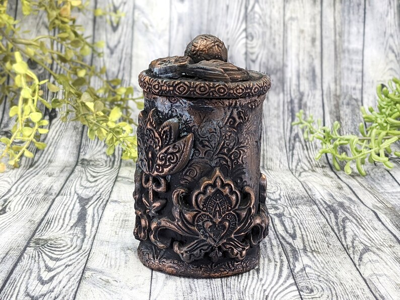 May include: A small, round, decorative container with a lid. The container is made of a dark brown material and has a textured, floral design. The lid is also decorated with a floral design.