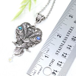 May include: Silver pendant necklace featuring two intertwined ravens with blue and green gemstone eyes. The pendant is approximately 1 1/2 inches tall and is measured against a ruler.