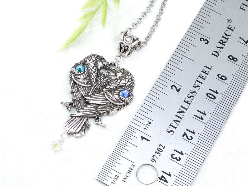 May include: Silver pendant necklace featuring two intertwined ravens with blue and green gemstone eyes. The pendant is approximately 1 1/2 inches tall and is measured against a ruler.