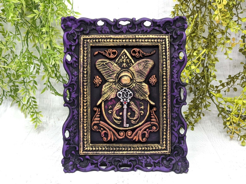 May include: A purple ornate frame with gold accents surrounds a black background with a gold butterfly and key design. The butterfly has a large eye in the center and the key has a red jewel.
