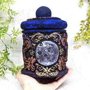 May include: A decorative ceramic jar with a blue and gold design. The jar has a lid with a moon-shaped design and a silver finish. The jar is decorated with intricate floral patterns.