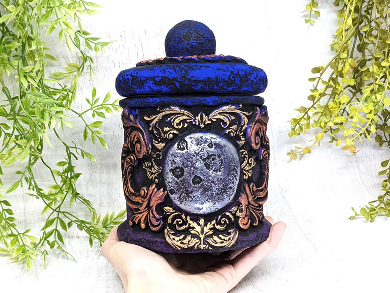 May include: A decorative ceramic jar with a blue and gold design. The jar has a lid with a moon-shaped design and a silver finish. The jar is decorated with intricate floral patterns.