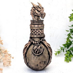 May include: A decorative bottle featuring a sculpted dragon head stopper, a textured bronze-toned finish, and a circular pendant depicting a claw. The bottle neck has a lattice pattern, and the rounded body is adorned with wing-like details.