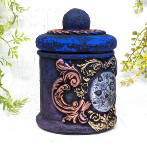 May include: A decorative ceramic jar with a blue and purple painted finish. The jar has a lid with a gold and copper design and a silver moon with a textured surface.