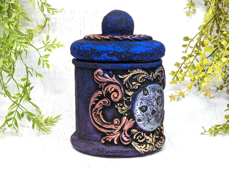May include: A decorative ceramic jar with a blue and purple painted finish. The jar has a lid with a gold and copper design and a silver moon with a textured surface.