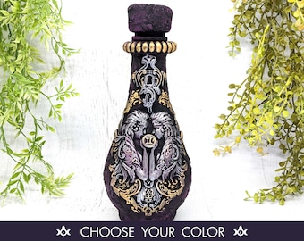 May include: Purple glass bottle with a silver and gold design featuring the Gemini zodiac symbol. The bottle has a textured surface and a decorative stopper.