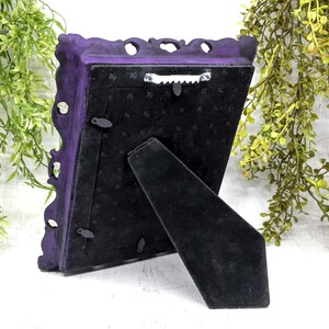 May include: A purple ornate picture frame with a black velvet backing and a kickstand. The frame has a decorative design and is made of metal.