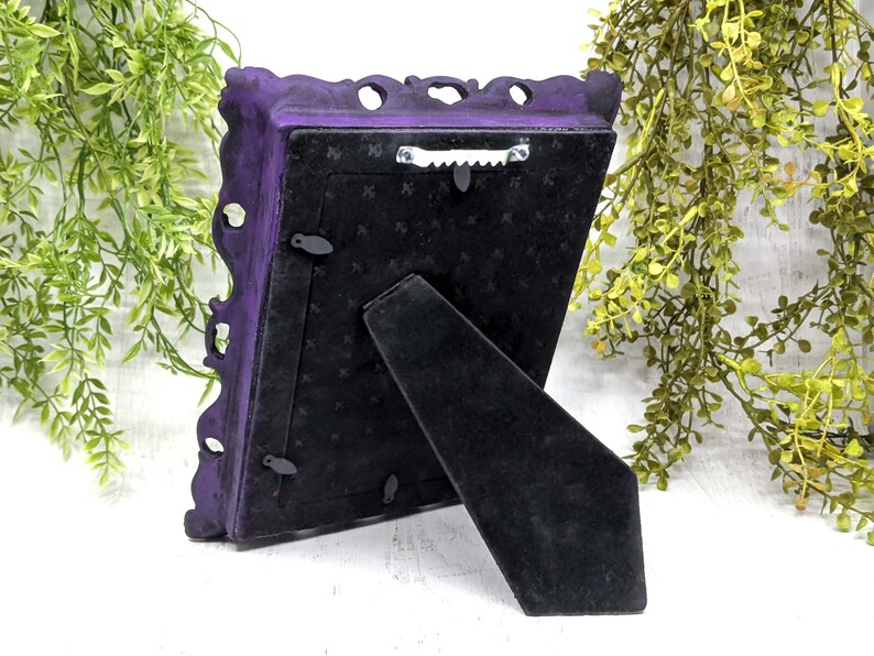 May include: A purple ornate picture frame with a black velvet backing and a kickstand. The frame has a decorative design and is made of metal.