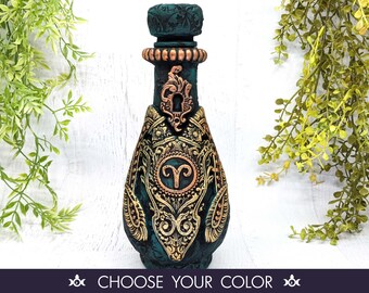 May include: A dark green glass bottle with gold accents and an ornate design. The bottle features a gold zodiac symbol of Aries. The bottle is decorated with a keyhole design.