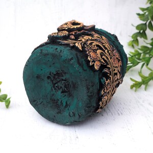 May include: A small, teal green planter with gold floral details. The planter has a distressed finish and is decorated with a gold floral pattern.