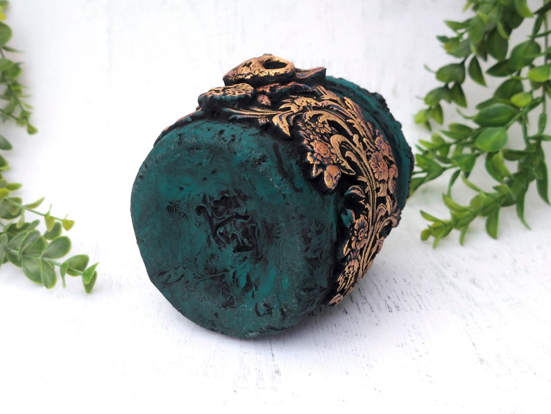 May include: A small, teal green planter with gold floral details. The planter has a distressed finish and is decorated with a gold floral pattern.