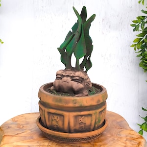 May include: A terracotta pot with a green leafy plant growing out of a brown, sculpted face. The pot has a decorative design and is sitting on a wooden surface.