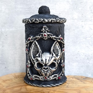 May include: A black decorative jar with a lid, featuring a bat skull design. The jar has silver accents and red gems, with a textured surface. The jar is cylindrical and sits on a wooden surface.