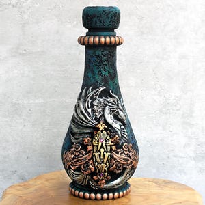 May include: A teal decorative bottle with a detailed dragon design in silver, gold, and copper tones. The bottle has a textured surface and a decorative stopper. The bottle is displayed on a wooden surface.
