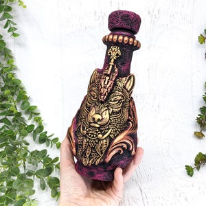 May include: A ceramic bottle with a detailed wolf design in gold and a burgundy base. The bottle has a decorative stopper with a keyhole design.