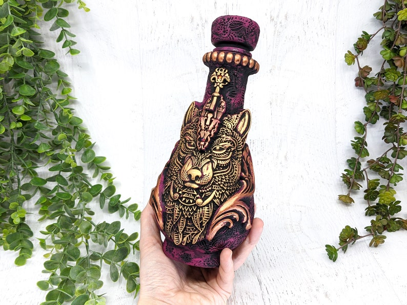 May include: A ceramic bottle with a detailed wolf design in gold and a burgundy base. The bottle has a decorative stopper with a keyhole design.
