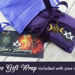 May include: A purple gift bag with a satin ribbon and a small charm with a triple moon symbol and a pentagram. The bag is sitting on a wooden surface. The text "Lori's Laboratory charm your spirit, craft your intention" is on a card in the background. The text "Free Gift Wrap included with your order!" is at the bottom of the image.