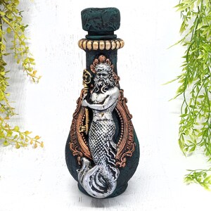 May include: A green ceramic bottle with a gold and silver mermaid design. The mermaid is holding a key and has a beard. The bottle has a gold rim and a gold and silver design around the neck.