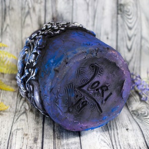 May include: A dark blue ceramic planter with a detailed floral design and a purple and black base. The planter has a signature on the bottom that reads "Yo!"