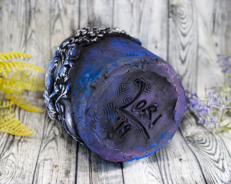May include: A dark blue ceramic planter with a detailed floral design and a purple and black base. The planter has a signature on the bottom that reads "Yo!"