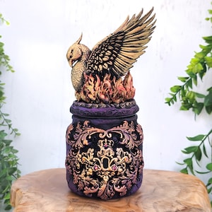 Könnte beinhalten: Eine dekorative Dose in Lila und Gold mit einer Phönix-Figur auf dem Deckel. Der Phönix ist aus Gold und hat seine Flügel ausgebreitet. Die Dose hat ein detailliertes Design mit goldenen Akzenten.
