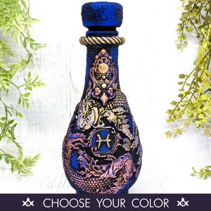 May include: A blue glass bottle with a gold and purple design. The bottle features a zodiac symbol for Pisces and a decorative stopper.