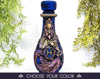 May include: A blue glass bottle with a gold and purple design. The bottle features a zodiac symbol for Pisces and a decorative stopper.