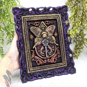 May include: A purple ornate frame with gold accents surrounds a black background with a gold butterfly and key design. The butterfly has a large eye in the center and the key has a red jewel.