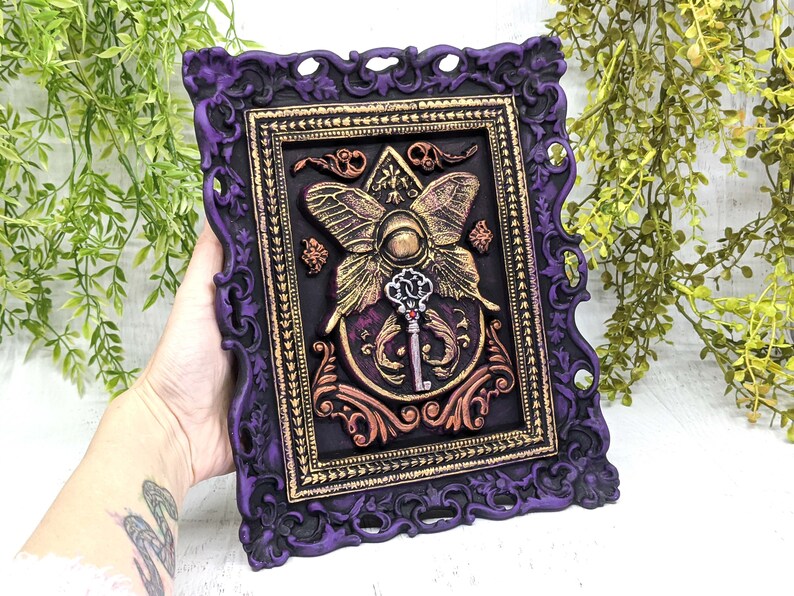 May include: A purple ornate frame with gold accents surrounds a black background with a gold butterfly and key design. The butterfly has a large eye in the center and the key has a red jewel.