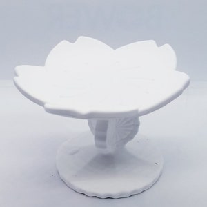 Op de afbeelding: Een witte keramische voetenschaal met een bloemvormige bovenkant. De schaal heeft een ronde basis en een steel met een decoratief ontwerp. Het ontwerp is eenvoudig en elegant, geschikt voor het presenteren van kleine items.