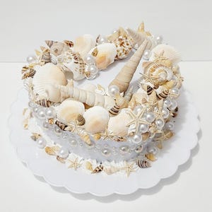 May include: A white cake decorated with seashells and pearls. The cake is on a white cake stand.