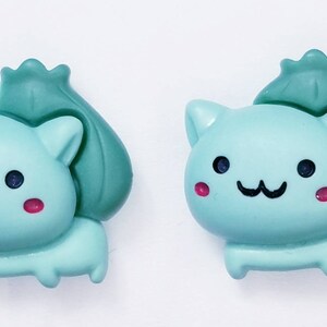 May include: Two light blue cat-shaped figurines with a green sack on their backs. The cats have black eyes, pink cheeks, and black smiles. These decorative figurines are perfect for cat lovers.