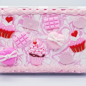 May include: Pink and white marbled box with a pink frosting border. The box is decorated with pink and red cupcake, chocolate bar, heart, and bow designs.