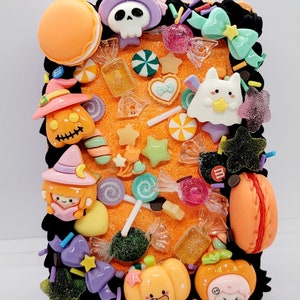 Puede incluir: Una funda para teléfono con temática de Halloween con un borde negro y un fondo naranja. La funda está decorada con varios encantos con temática de Halloween, incluyendo una calavera, un fantasma, una bruja, calabazas, caramelos y estrellas.