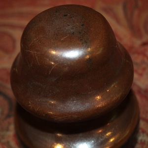May include: A vintage, copper-toned bottle stopper with a rounded top and a flared base. The stopper has a smooth, reflective surface with visible wear and patina, suggesting age and use. The design is simple and functional.
