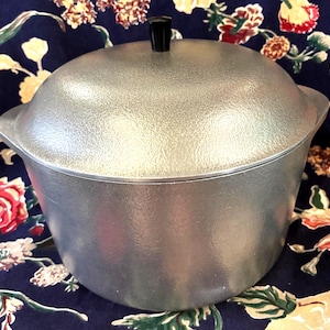 May include: A silver-colored metal pot with a textured finish and a matching domed lid. The lid features a small, black, cylindrical knob at the center. The pot has a wide, rounded body with integrated side handles, presenting a sturdy, utilitarian design.
