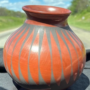 May include: A handcrafted, round, terracotta-colored pottery vase with a wide opening. The vase features a design of vertical, gray, teardrop-shaped patterns. The rim and interior are a darker shade of red. The vase is sitting on a black surface.