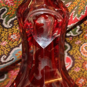 May include: A red glass bottle with a silver top and a decorative etched design. The bottle is sitting on a patterned fabric.