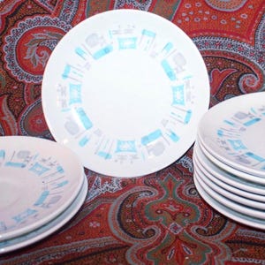 May include: A collection of white ceramic plates with a light blue and gray pattern. The plates feature a repeating design of stylized kitchen utensils and appliances. The image shows several plates stacked and arranged on a patterned fabric.