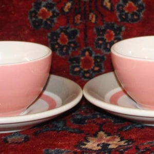 May include: Four pink teacups with matching saucers. The cups have a rounded shape with a handle, and the saucers feature a white and gray striped design. The interior of the cups is white, creating a contrast with the pink exterior.