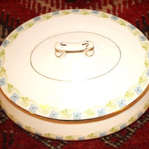 May include: An oval ceramic tureen with a lid and two side handles. The white surface features a decorative border pattern of light green swirls and small blue squares, accented by thin gold trim along the edges and the central lid handle.