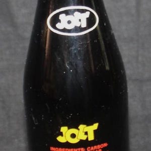 May include: A vintage Jolt Cola bottle with a brown cap. The dark glass bottle features the Jolt logo and text detailing ingredients and redemption value. The label reads "Populating The World."