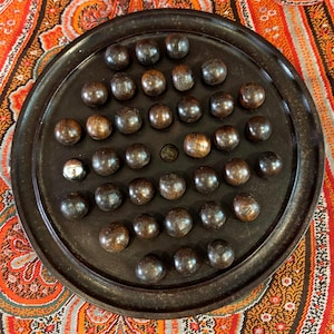 May include: A circular dark brown wooden board game featuring a grid of small indentations filled with numerous smooth, spherical dark brown wooden marbles, with one single metallic silver marble positioned near the left side of the board.