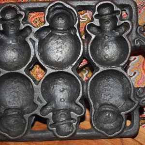 May include: A black cast iron baking mold featuring six individual cavities shaped like snowmen. The mold has two side handles and a rectangular frame. Each snowman cavity displays distinct details including hats, buttons, and star patterns.