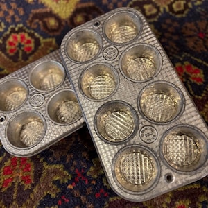 May include: Two silver-toned metal muffin pans with a textured grid pattern on the bottom of each cup. Each pan features circular indentations and is stamped with the text Ekco Chicago. The pans have a metallic finish and a rectangular shape.