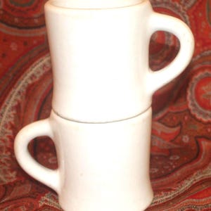 May include: Two white ceramic mugs stacked on top of each other. Each mug has a curved handle and a slightly flared shape. The mugs are set against a red and orange paisley patterned background.