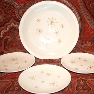 May include: A set of vintage white ceramic dishes with a gold starburst pattern. The set includes a large serving bowl and three smaller plates. The dishes are arranged on a red and brown patterned surface.